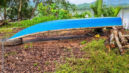 Konstfotografi Inverted Blue Boat on Shore: A vibrant blue boat rests upside down on a grassy shore, supported by logs and a tire, hinting at a rustic, coastal scene