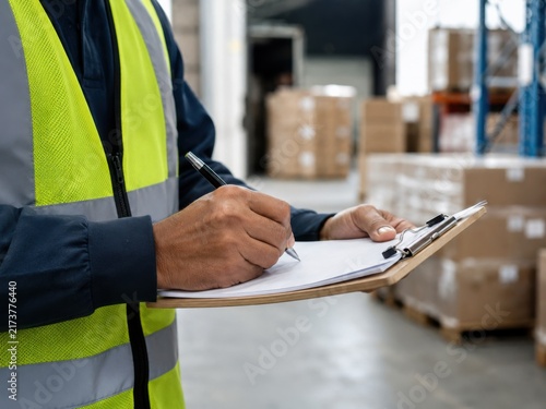 A worker in a high-visibility vest records inventory details on a clipboard inside a warehouse.