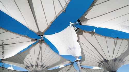 Large white fabric canopy structures stretch skyward against clear blue sky. These modern architectural designs offer shade, create geometric pattern with sunlit fabric panels, suspended supports.