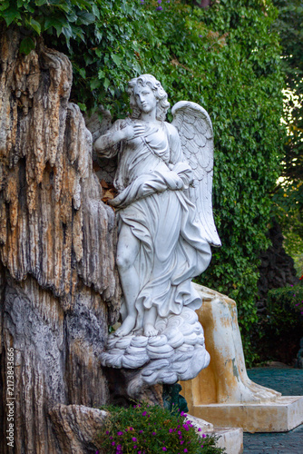 Fotografie White Angel Statue Near Rock Grotto and Ivy Wall, Medium Shot