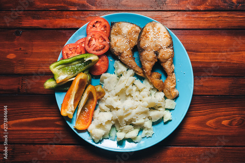 Pieces of fish fried in vegetable oil sit on a white blue plate next to mashed potatoes, tomato, and bell pepper (paprika). A perfect dish for breakfast, lunch, or dinner.
