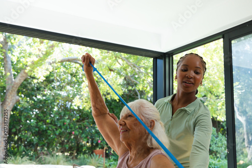 Obraz na plátne Senior woman lifting blue band African American female caregiver guiding in gree