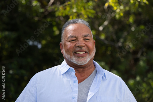 Senior African American man smiling, standing at park wearing light-blue shirt and gray T-shirt