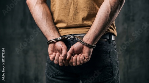 Rear view close-up of a man wearing a brown shirt and jeans with his hands handcuffed behind his back against a dark textured wall, representing arrest and legal justice.