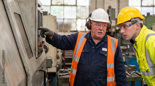 Senior factory technician teaching a trainee to inspect an industrial control panel. Worker in hard hat and hi-vis vest explaining machine operation to an apprentice in a manufacturing plant