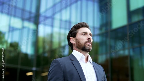 Serious mature businessman walks outside office building, standing in close up. Professional in formal suit confidently looking around with a composed, accomplished expression, success, determination.