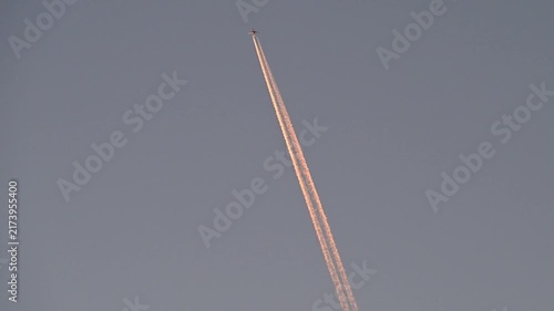 A commercial airplane high in the sky leaves long, golden sunset contrails as it flies through the evening air.