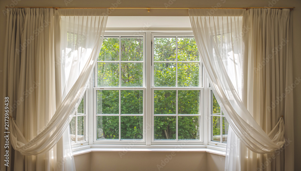 Fototapeta premium Bright window with sheer curtains framing a lush green garden view
