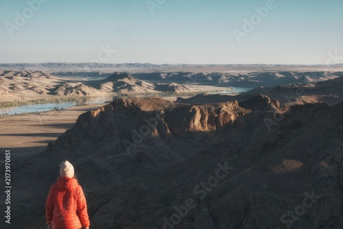 Photography A hiker in an orange jacket looks out over a rocky canyon and the winding Ili River in Kazakhstan, ideal for active lifestyle branding, adventure travel promotions, and ecotourism layouts