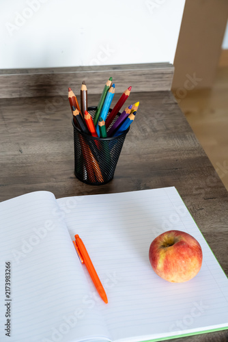 Fotografía Open blank notebook with orange pen, colored pencils, and red apple on wooden de