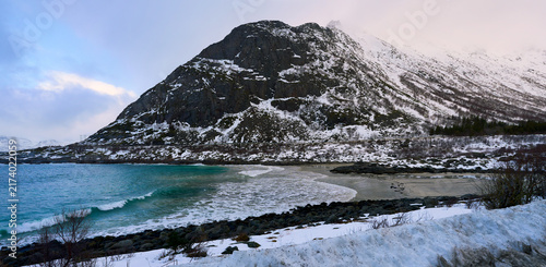 Beautiful scenic viewpoint of a coastal highway winding next to the blue sea in Lofoten, Norway. Majestic winter landscape with grand snow-covered mountains under a dramatic overcast sky in Arctic Sca