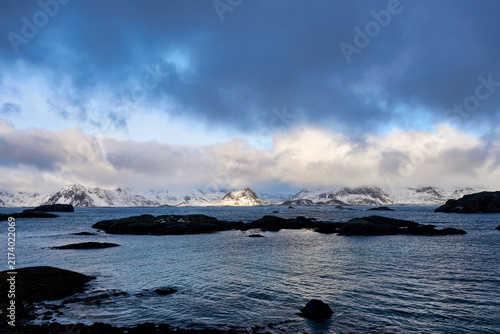 Beautiful scenic view of a traditional Norwegian fishing village with red wooden cabins by the fjord in Lofoten, Norway. Majestic snow-covered mountains under a dramatic winter sky in Arctic Scandinav