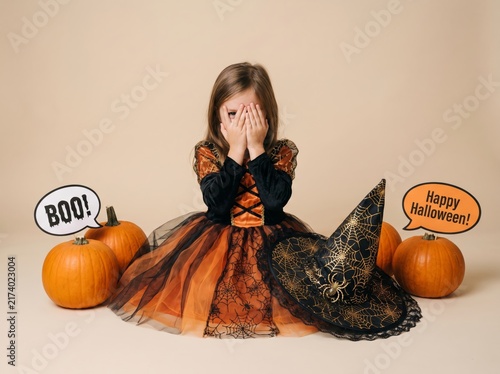 Little girl in a festive black and orange Halloween witch costume covering her face playfully surrounded by pumpkins and a witch hat on a light studio background celebrating autumn holiday