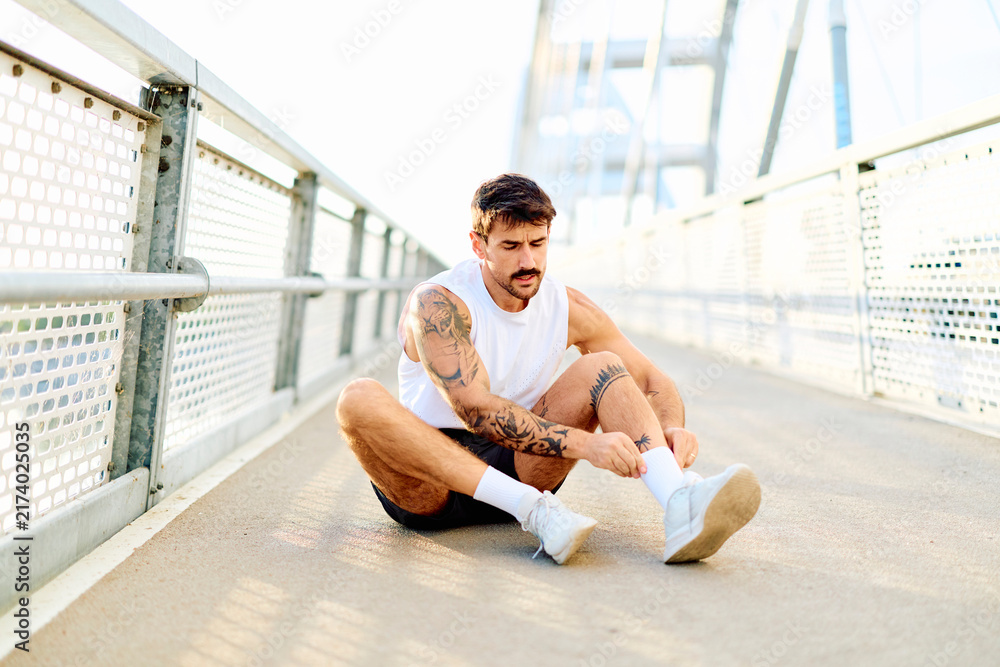 Obraz premium Young athletic man with tattoos resting on a bridge, adjusting socks and preparing for or recovering from a run or exercise