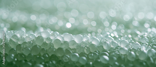 Macro close-up of numerous translucent, sphere-like gel beads or textured plastic pellets with a sparkling, softly blurred green bokeh background, creating a serene and abstract shimmering