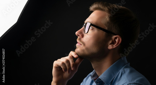 Thoughtful young man in glasses pondering against a dark background
