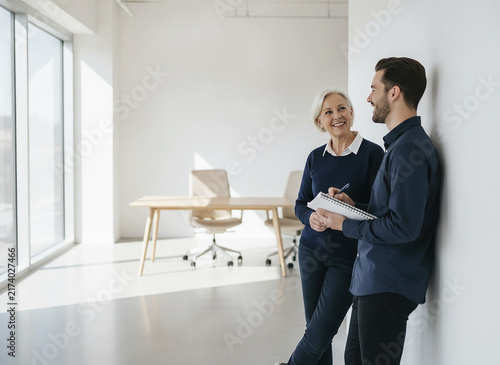 Business colleagues talking near wall, casual workplace interaction. Senior woman and young man having a conversation in a bright office. Professional team members collaborating on a project in