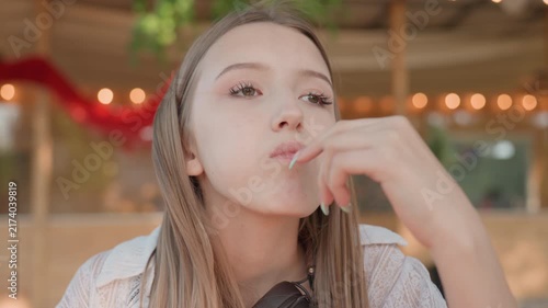 Close up of young woman with long straight hair wearing white lace top and headphones making expressive hand gestures including pointing finger upwards. Background features warm bokeh lights