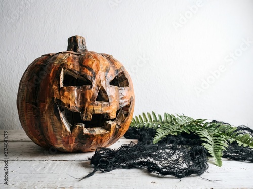 Distressed carved Halloween pumpkin jackolantern with spooky face resting on a rustic white wooden surface alongside green fern and black cobweb decorations autumn season
