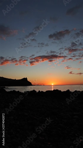 Stunning vertical sunrise over the calm Atlantic Ocean in Madeira, featuring scenic dark rocky coastline silhouettes under dramatic orange clouds at dawn. 4K vertical video.