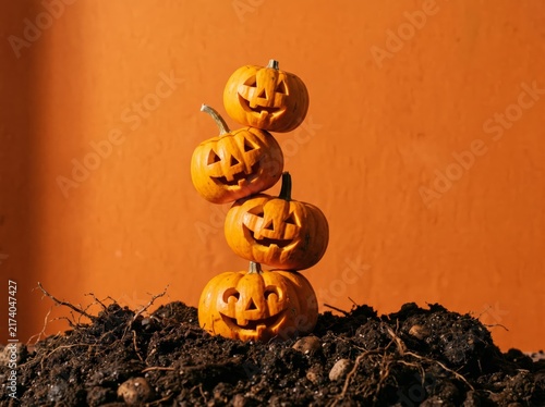 Four smiling carved Halloween pumpkins stacked vertically on a dark soil pile against a vibrant orange background creating a festive autumn display with shadows