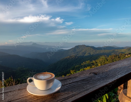 Latte coffee cup on wooden table with mountain landscape background at sunrise.