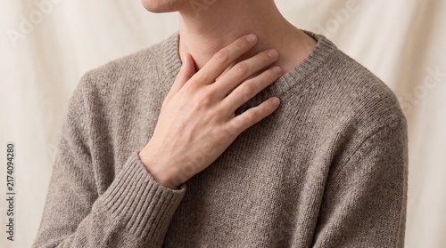 Person wearing brown knitted sweater resting hand gently near throat against neutral soft fabric background with captures close view