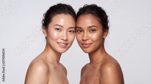 Diverse skin tones two women with glowing complexions side by side smiling gently with posed closely together against neutral background
