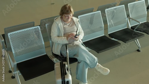 Mature passenger sits among rows of empty seats in airport terminal hall. Senior woman waits for boarding and uses smartphone. Quiet departure gate area offers seating during flight delay.