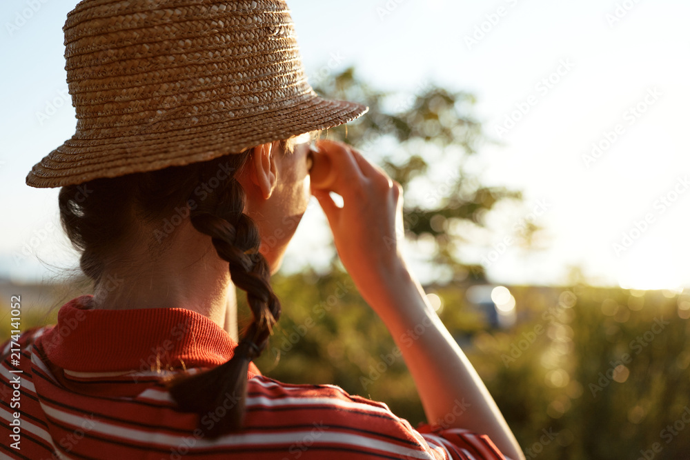 Naklejka premium Young woman in a straw hat with braided hair looks through binoculars at the sunny meadow. An authentic lifestyle scene captures a nature adventure and exploration.
