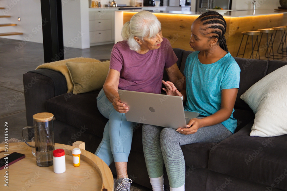 Naklejka premium Senior woman and African American woman sitting on sofa at home using laptop with pill bottles