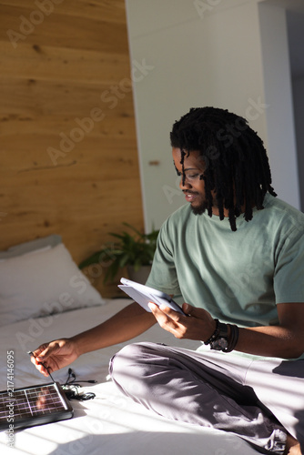 Man sitting cross-legged on made bed in bedroom with wooden wall and diagonal sunlight