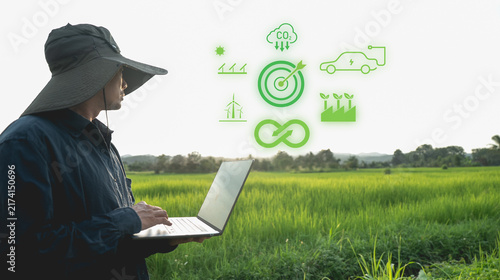 Person using laptop in lush green field with eco friendly icons floating above, symbolizing sustainable technology and renewable energy