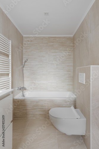 Minimalist modern bathroom interior with bathtub, wall mounted shower, beige textured tile, wall hung toilet, bright neutral finishes, clean residential layout, and refined spa inspired comfort