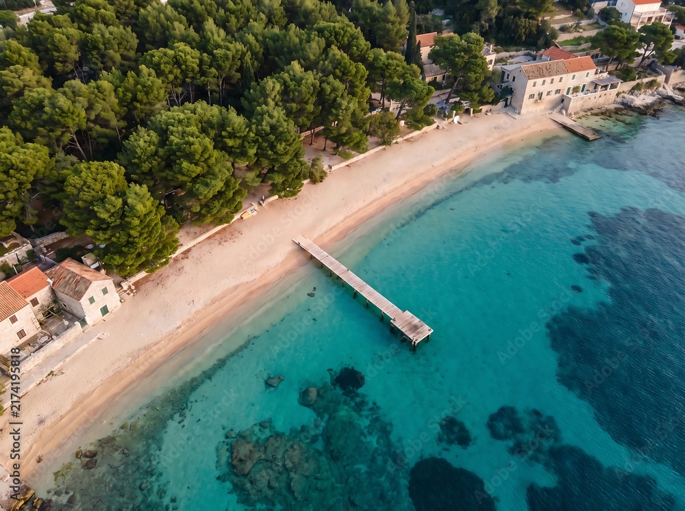 Fototapeta premium Aerial view of a scenic coastal beach with crystal clear turquoise water a wooden pier green pine trees along the shoreline landscape tropical