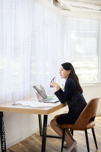 Young Japanese businesswoman holding her glasses and smiling while looking out of the window at a desk
