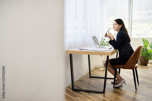 Young Japanese businesswoman holding her glasses and smiling while looking out of the window at a desk