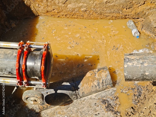 Fotografie Hydraulic butt fusion welding machine and severed black HDPE pipe in a flooded muddy trench during emergency water main repair