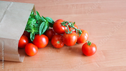 Fresh tomatoes and basil spill out of a brown paper bag on a wooden table in a kitchen setting