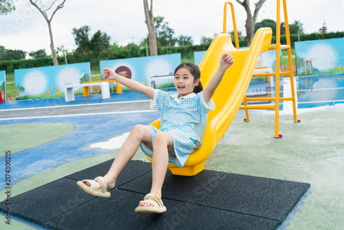 Happy young girl enjoying sliding down yellow playground slide outdoor in school uniform on soft safety mat surface