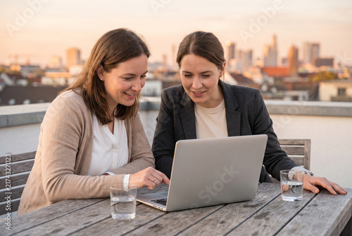 Women working together on a laptop while sitting outdoors at a table with city view
