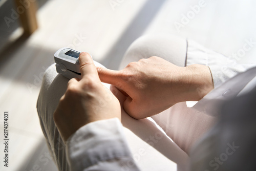 Obraz na płótnie Person Measuring Oxygen Saturation With Fingertip Pulse Oximeter On Finger While Seated On Chair In Bright Indoor Setting With Sunlight On Lap White Clothing And Neutral Background