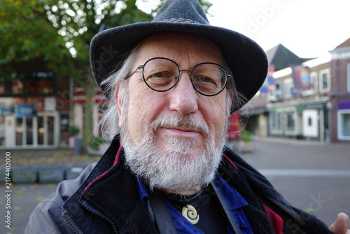 Senior man with a beard and glasses wears a dark felt hat. He poses outdoors in the Netherlands, against a backdrop of a tree and buildings featuring red brick and large windows. 