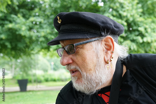 Older man wearing a black captain's hat with an anchor emblem, glasses, and multiple earrings. He has a white beard and is dressed in black clothing. 