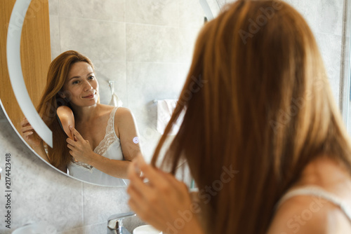 Foto Woman brushing long hair looking in bathroom mirror