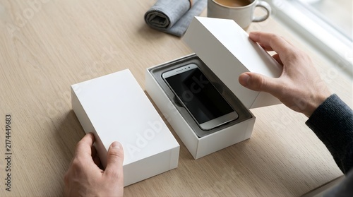Person opening a white box containing a new smartphone on a wooden desk