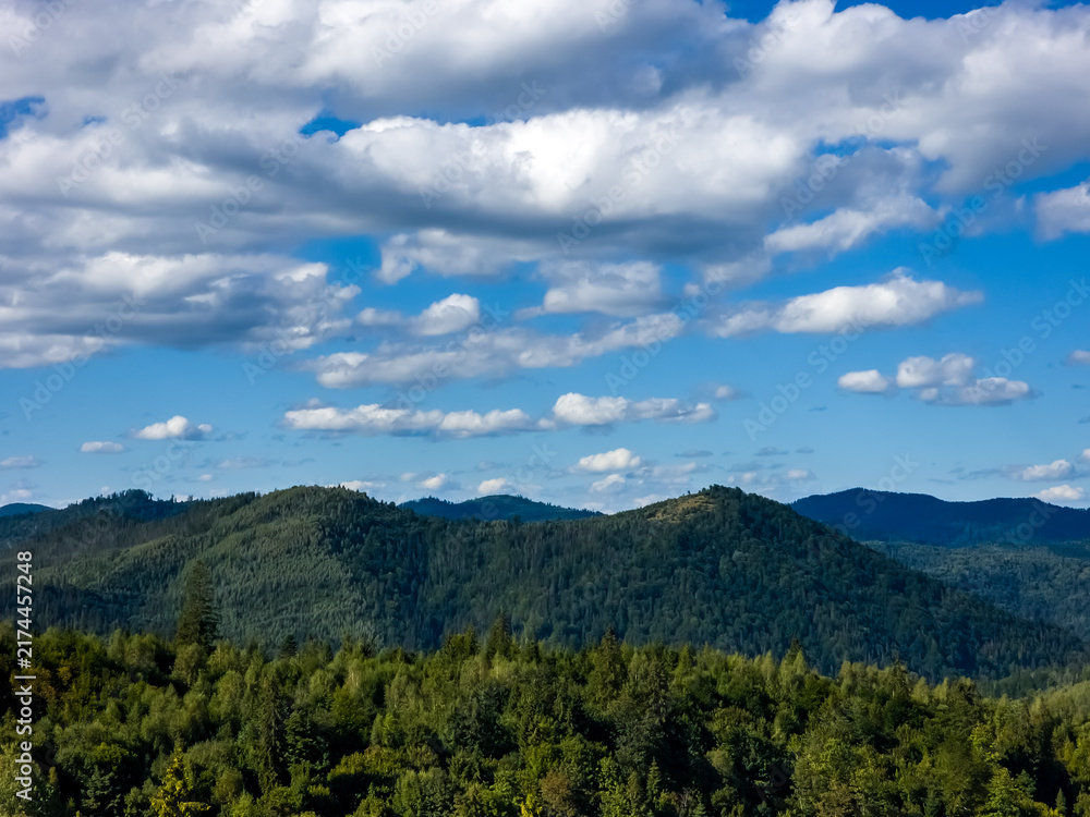 Fototapeta premium Panoramic view of green mountain forest range under bright blue sky with fluffy white clouds, natural background template for eco tourism and outdoor hiking adventure advertising banner