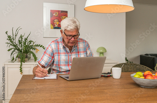 An elderly man with white hair sitting at a wooden dining table writing in a notebook