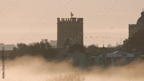 Silhouetted medieval castle tower with the ukrainian flag waving against a hazy sky as thick morning fog rolls through the city, creating a mystical and historical atmosphere
