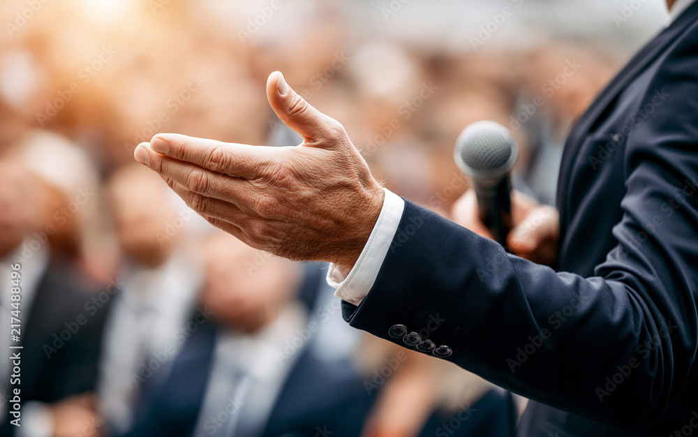 Fototapeta premium Public speaker hand gesturing during presentation in front of blurred audience